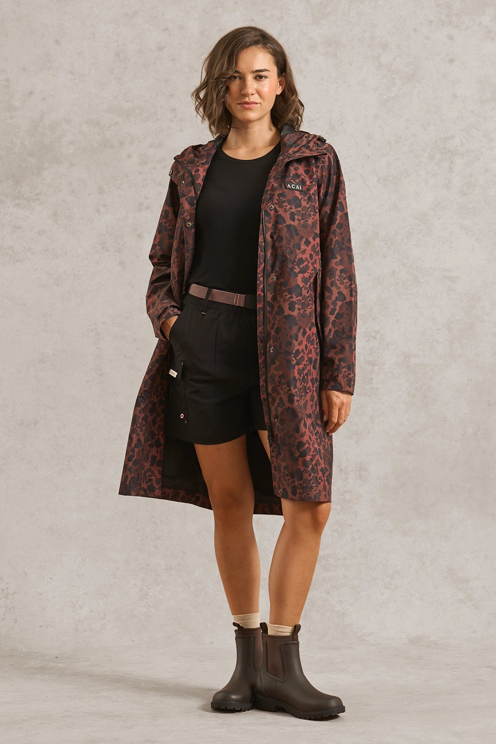 A woman models a mid-length hooded raincoat with a dark brown and black leopard print pattern, worn over a black outfit with brown Chelsea boots, against a textured gray studio backdrop.