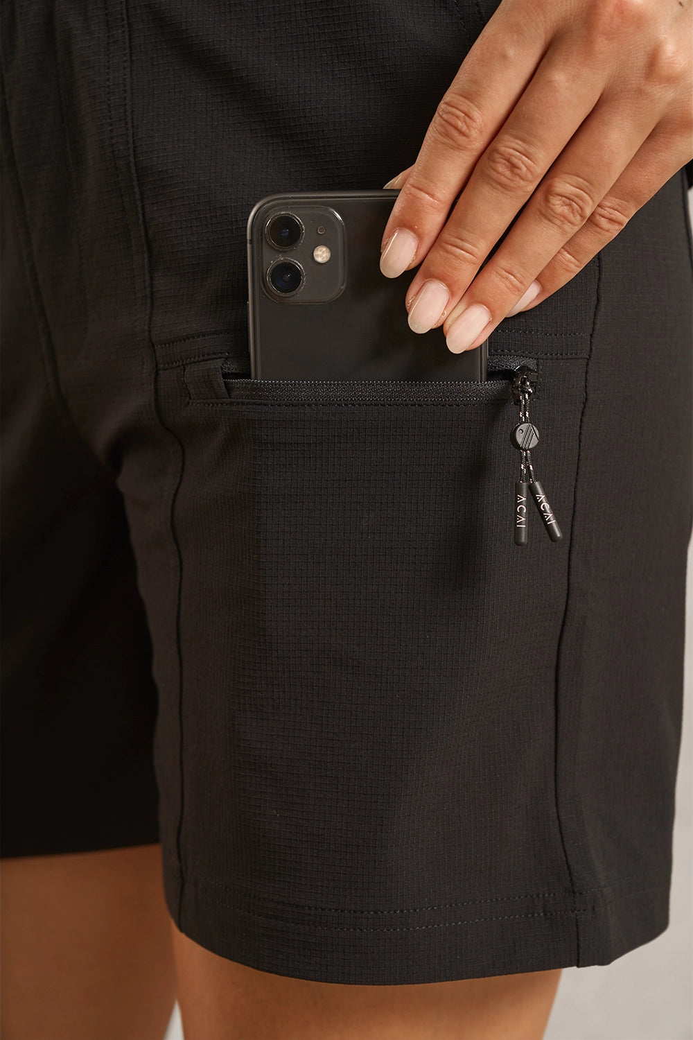 Close-up of a person wearing black textured shorts, showing a side pocket with a smartphone tucked into the pocket and a zipper pull hanging from the side.