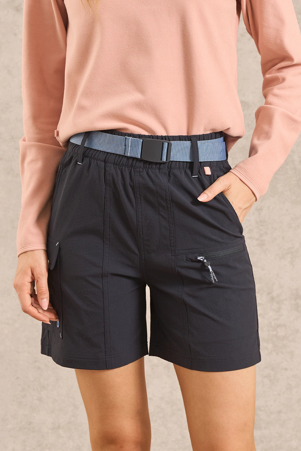 A person wearing dark navy cargo shorts with a blue belt, paired with a dusty pink long-sleeve top, shown from the chest down against a neutral textured background.
