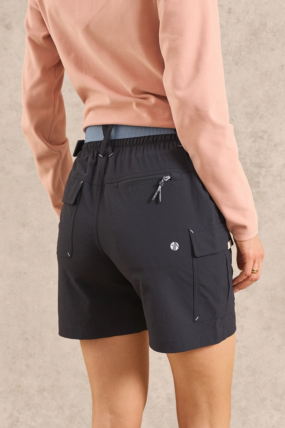 A person shown from the back wearing dark charcoal cargo shorts with zippered pockets and a peach long-sleeve top, standing against a textured beige wall.