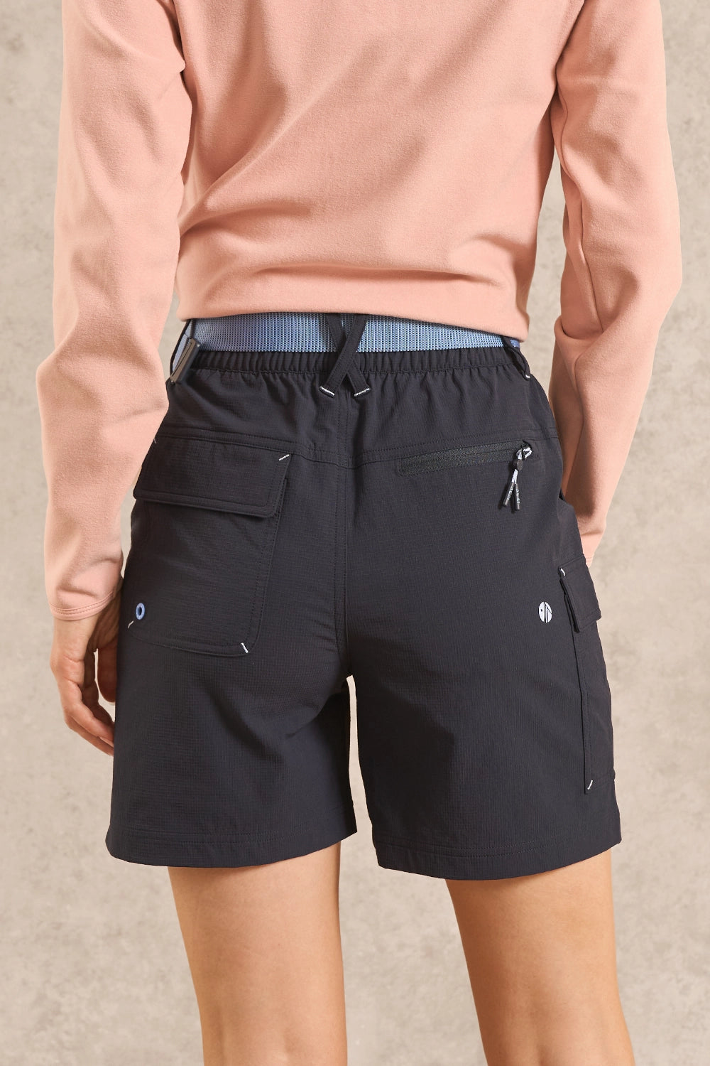 Rear view of a person wearing dark navy cargo shorts with a drawstring waist and side zippers, paired with a peach long-sleeve top against a neutral textured background.